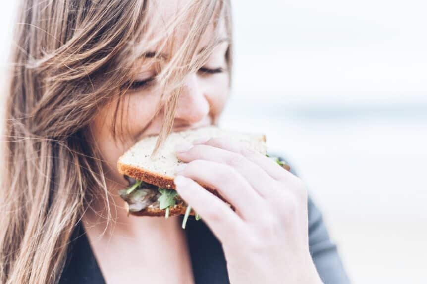 A woman with windswept hair biting into a sandwich, hosted by Wellin5